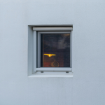 Una finestra fotografata dall'esterno che lascia intravedere una lampada accesa.