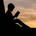 Bambino in ombra che legge un libro al tramonto