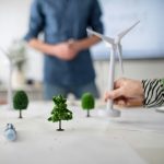 Mano che regge la miniatura di una turbina eolica e la poggia su un tavolo tra miniature di alberi
